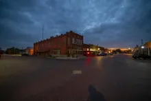 Two-story red-brick commercial building at a street intersection during evening twilight, with illuminated storefronts and a cloudy sky overhead.